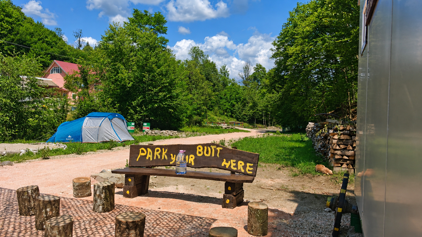 Banca de lemn din camping cu inscriptia Park your butt here, cu corturi si stiva de lemne in fundal